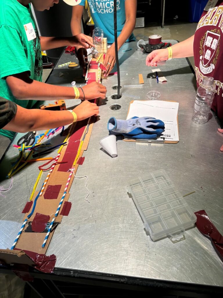 Four students surround a table. On the table is a cardboard runway edged with straws, beginning with a ramp.