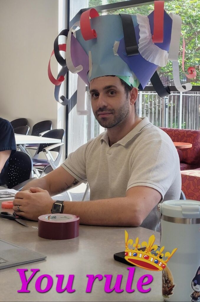 A man sits at a table with a very large paper hat on his head.  Text at the bottom says "You rule"